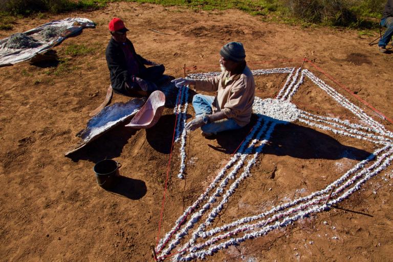 Two Aboriginal people sitting in the outback creating an image on the ground using white paint.