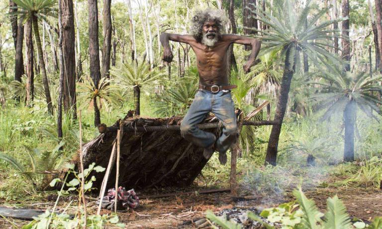 An older David Gulpilil jumping up in the air in the bush. He's in front of a lean-to shelter.