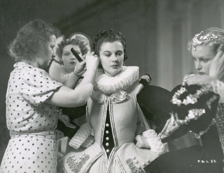 Vivien Leigh in costume on the set of Fire Over England has her hair adjusted by an assistant holding a comb as two other actors in costume look on