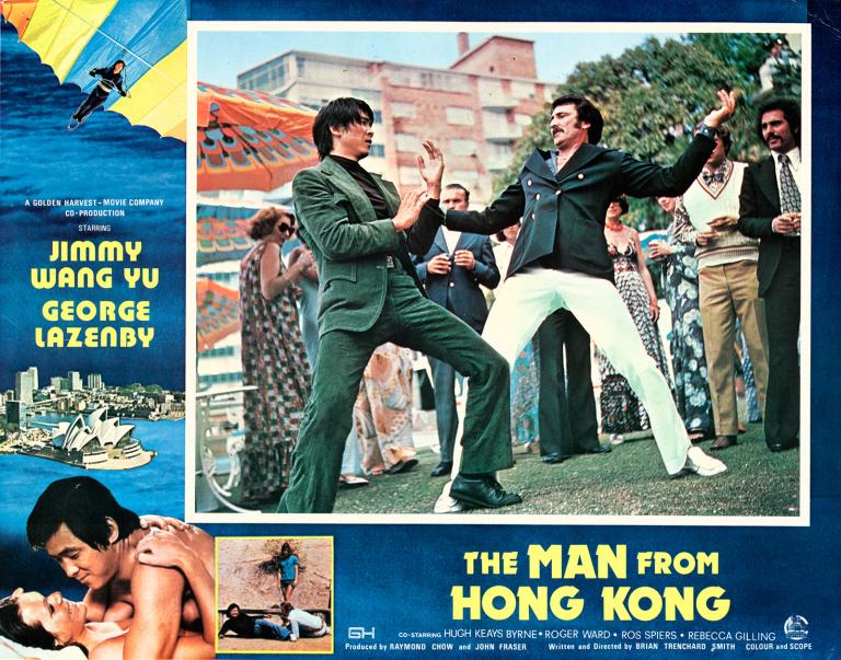 Lobby card with main image of 2 men, 1 in a dark grey suit, the other in white pants and dark jacket are fighting in a martial arts style surrounded by onlookers. Around this image is the film title on blue background and other scenes from the film.