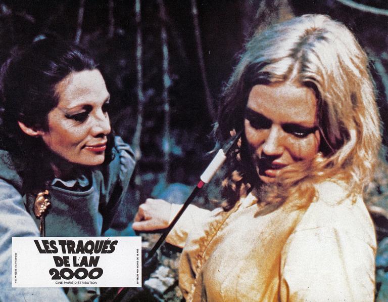 Close up of a menacing-looking dark-haired woman (Carmen Duncan) tormenting a blond woman (Lynda Stoner) by pointing an arrow at her face in a scene from 'Turkey Shoot'.