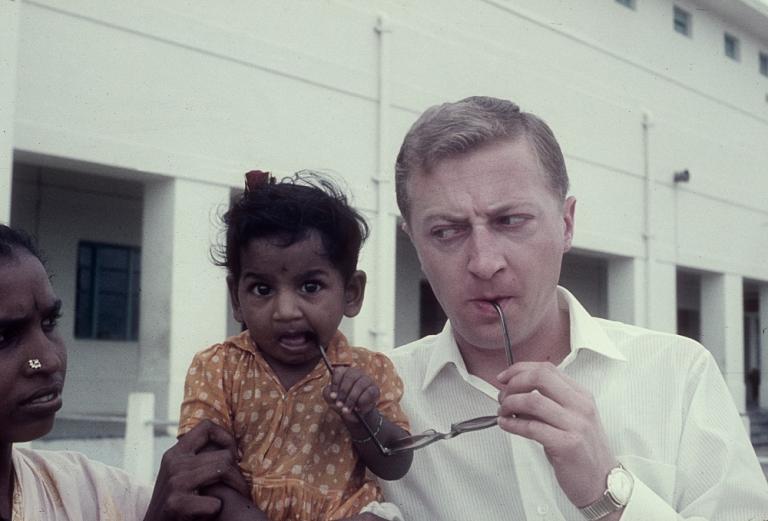 An Indian mother hold her child while the child and Graham Kennedy suck on the arms of Kennedy's glasses. Kennedy has a wry expression