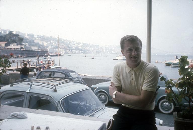 Graham Kennedy, arms folded, stands in front of cars at an Italian seaside town