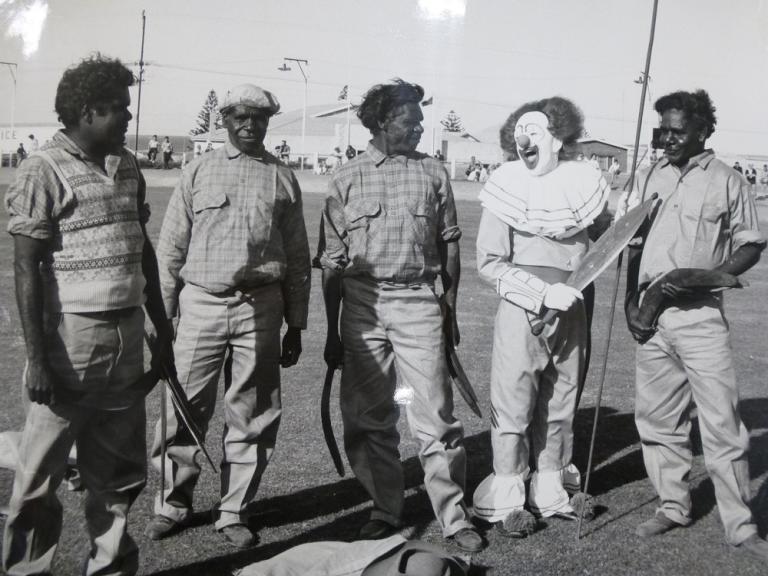 Bobo the Clown with a group of Aboriginal men holding spears, boomerangs and a woomera