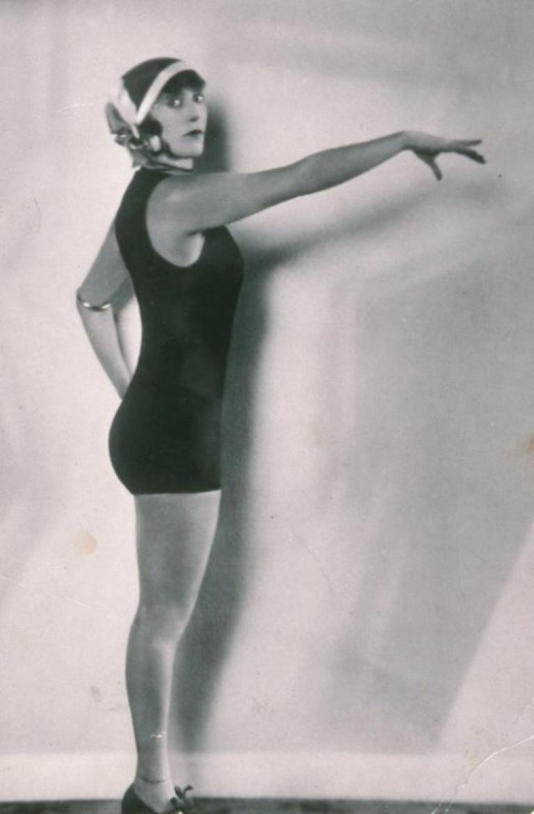 Black and white image of Annette Kellerman wearing a one-piece bathing suit and holding her arm out in front of her. She is looking at the camera.