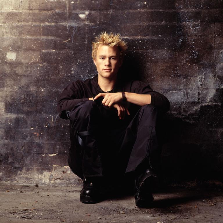 Heath Ledger crouches up against a wall looking at the camera. 