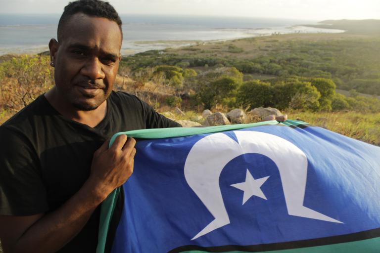 A Torres Strait Islander man holds the Torres Strait flag with the coast behind him