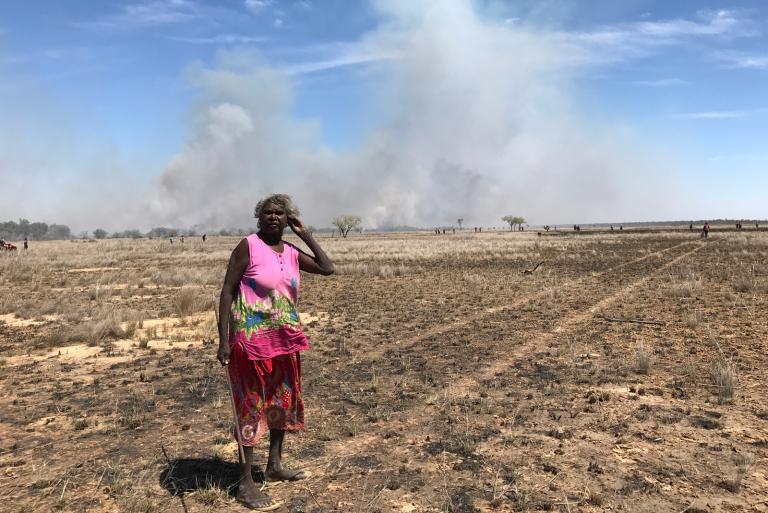 An older Aboriginal woman stands in the middle of a dusty plain in the outback.