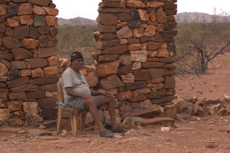 A man sits on a chair in front of a stone wall on a plain in the outback.
