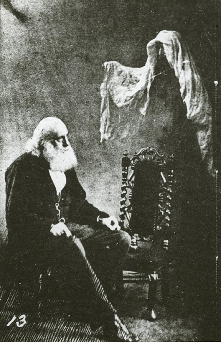 Glass sldie of an old, bearded man setaed with a ghost hovering to his left with its arm extended