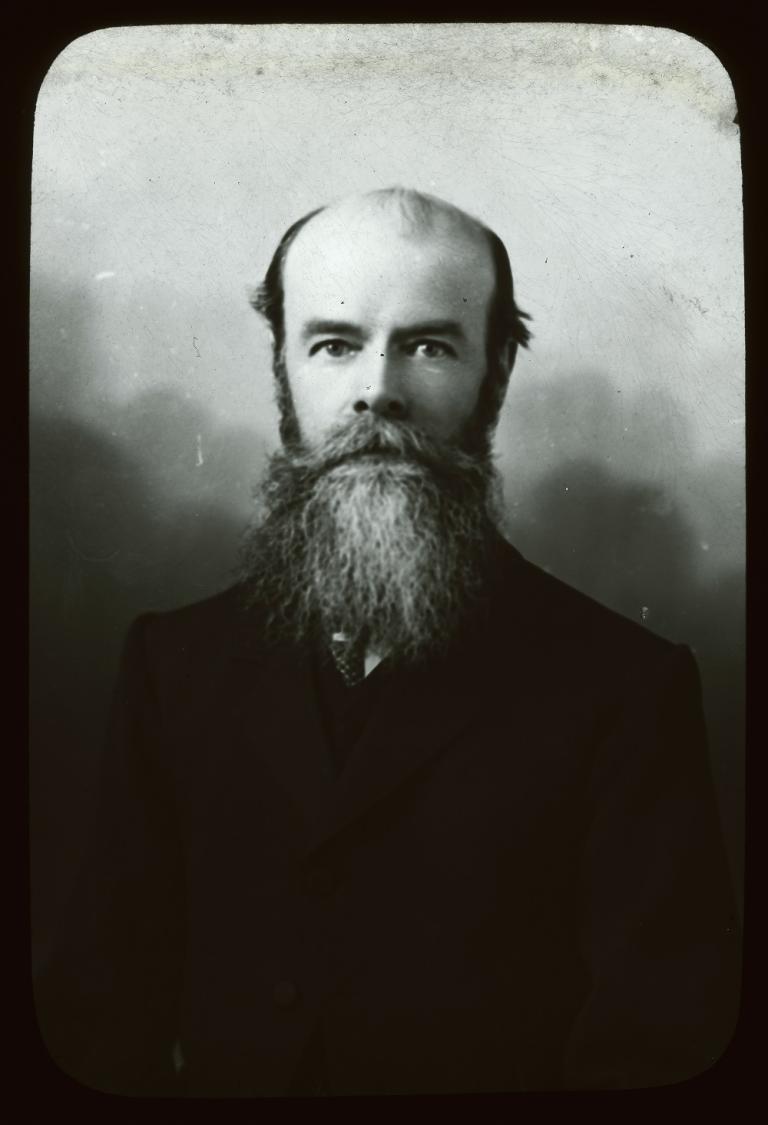 Black and white glass slide showing the upper body of a man with a dark moustache and beard, wearing a dark jacket, looking into the camera. 