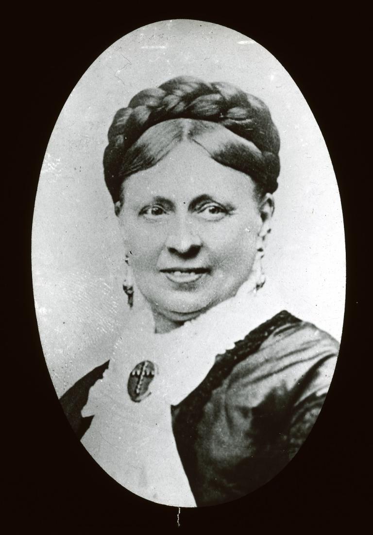 :  Black and white glass slide showing the head and shoulders portrait of a woman wearing a pale blouse with a brooch attached and a dark jacket, turning head to her left, looking left, smiling. 