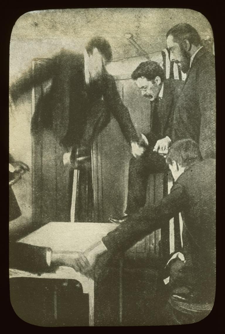 Sepia glass slide showing approximately five men and a spirit joining hands around a table, looking down at the table.