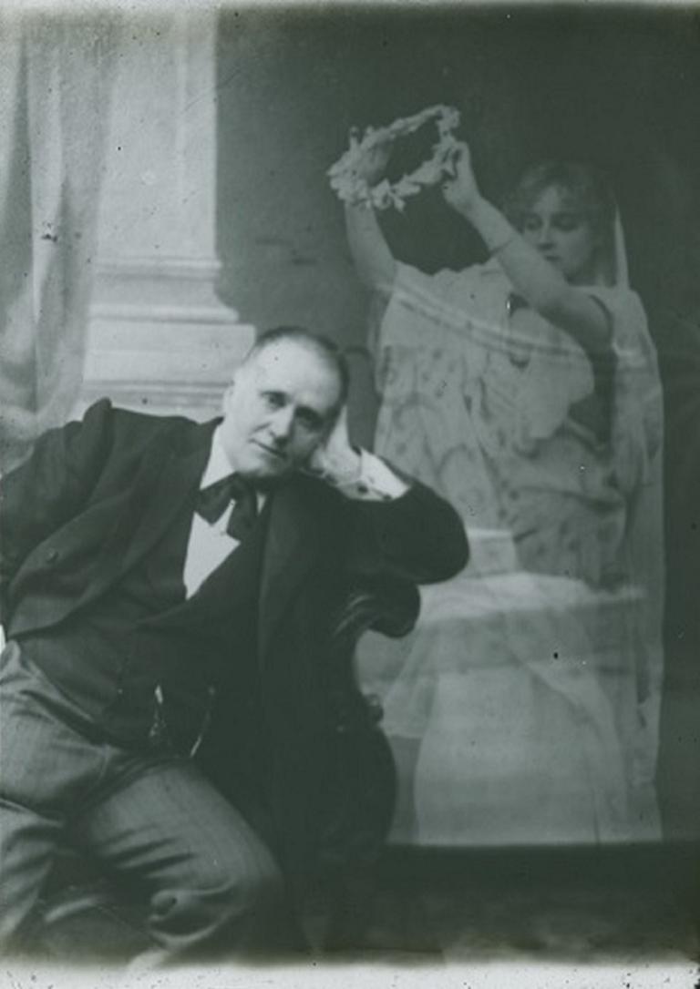 Black and white glass slide showing a man sitting on a chair resting his left arm over the side, a spirit hovering at his left holding a wreath over him.