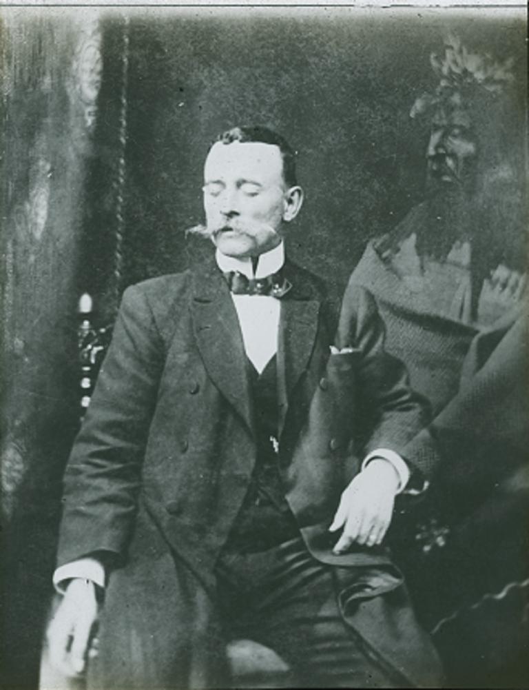 Black and white glass slide showing a man sitting on a chair with his eyes closed and a spirit partly superimposed over his left arm and chest.