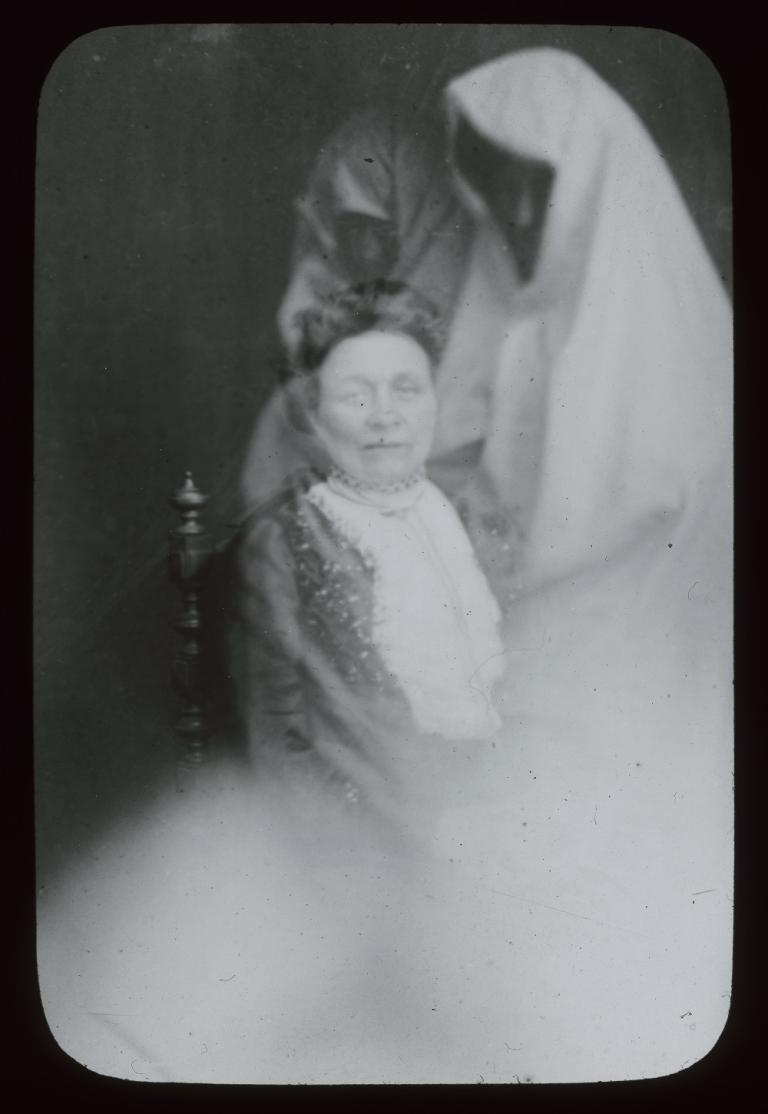 Black and white glass slide showing two spirits, one behind and one to the left of a woman sitting on a chair