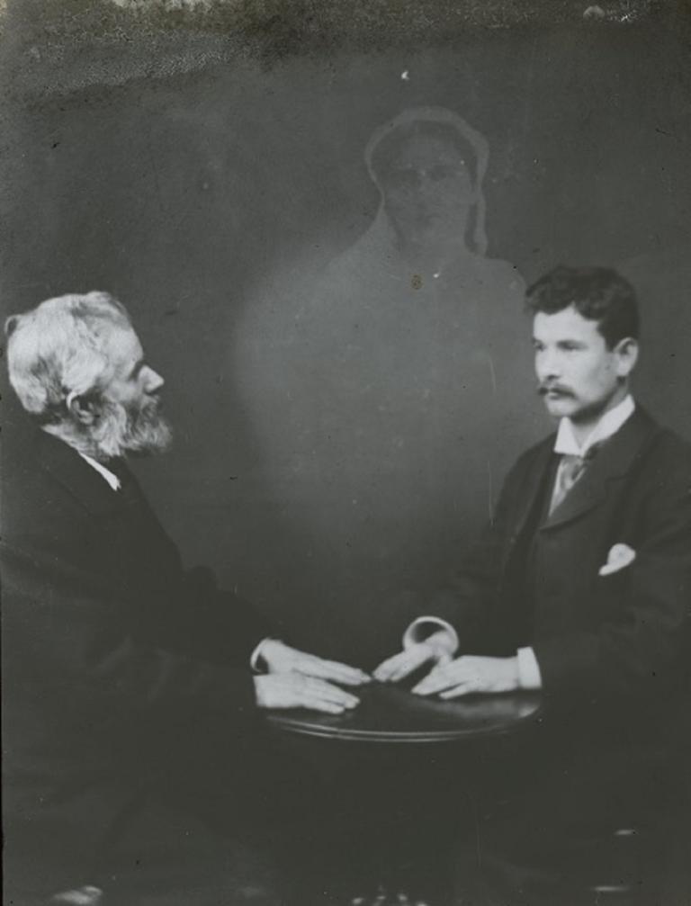 Black and white glass slide showing two men sitting opposite each other, hands resting on a table, a spirit hovering behind them.