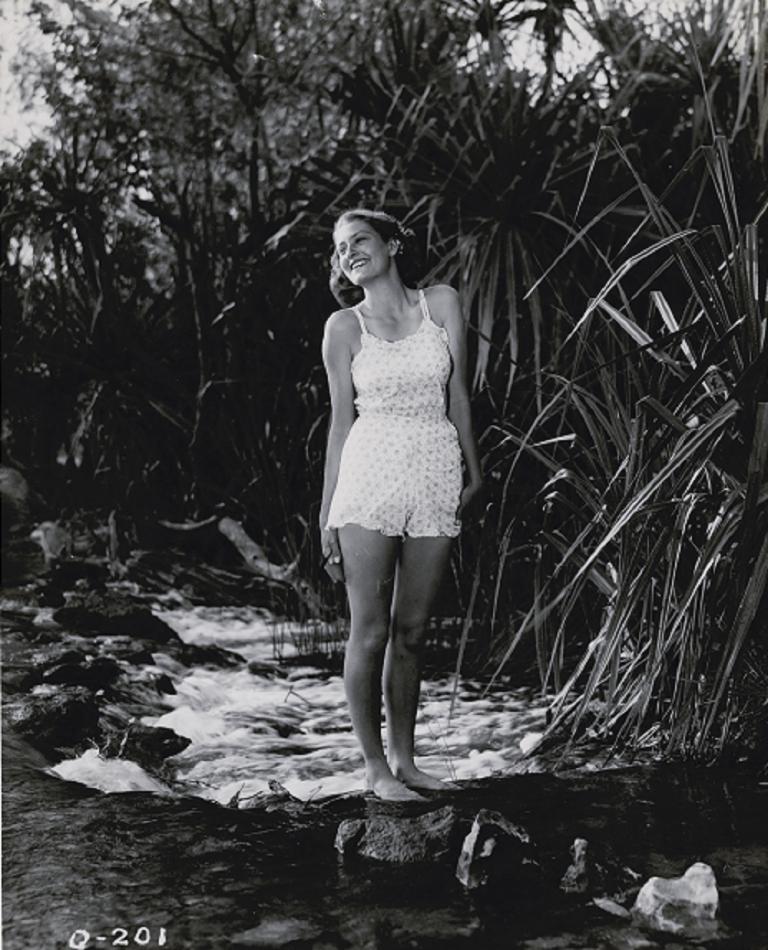 A woman wearing older style swimmers stands on a rock in the middle of a stream