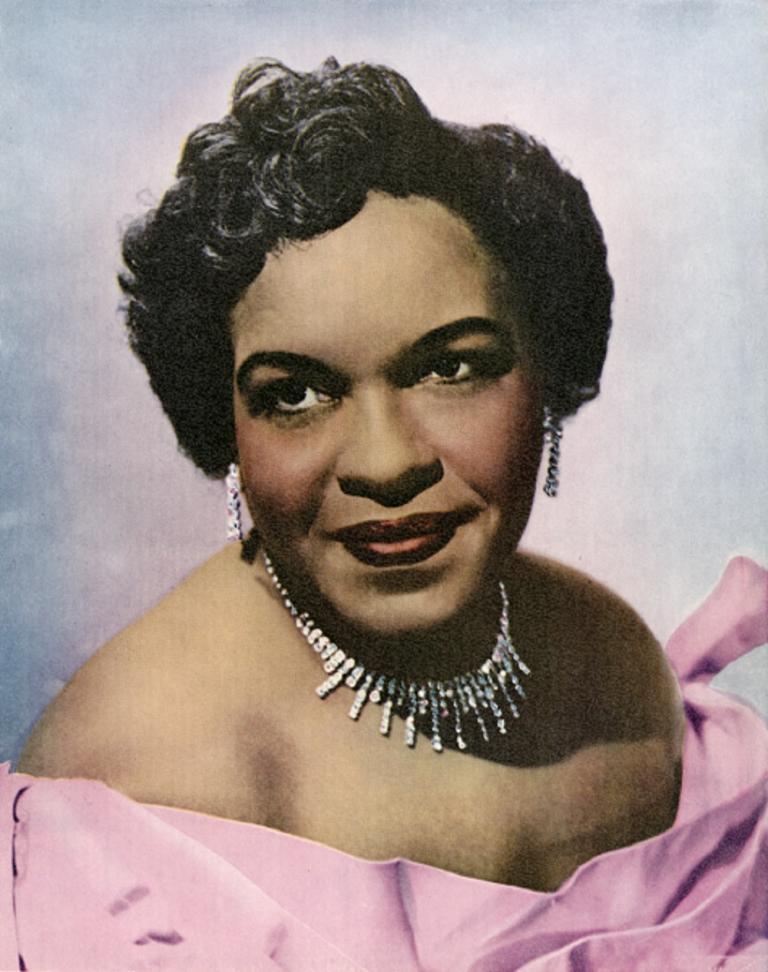 Coloured studio portrait of Winifred Atwell wearing a pink off the shoulder dress, ear rings and necklace
