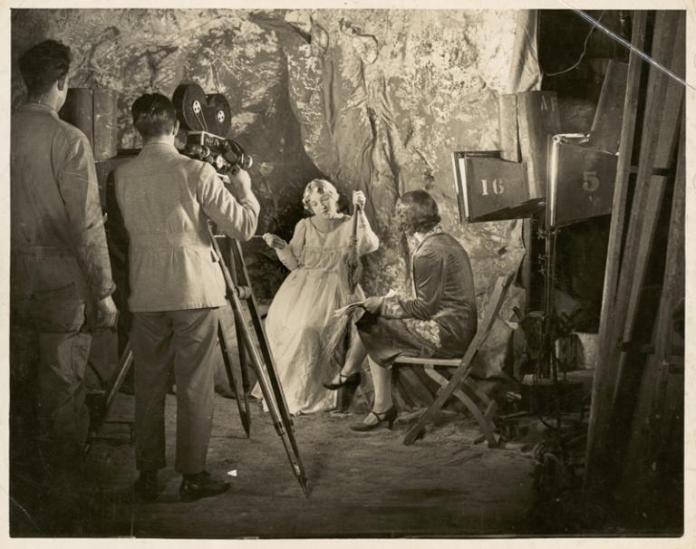A black and white photo of Paulette McDonagh directing an actor on-set. There is a two men in the forground. One of them is operating a film camera.