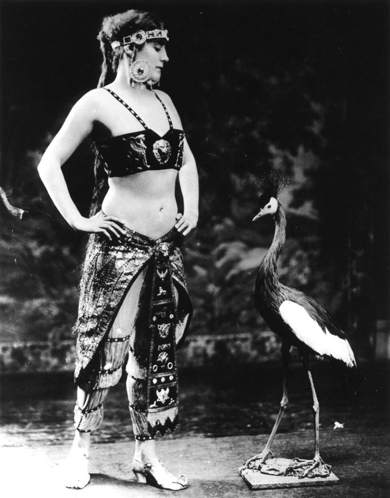 Actor Annette Kellerman stands with her hands on her hips, looking down at a taxidermied crowned crane bird. Annette is wearing a be-jewelled top, skirt and headdress.