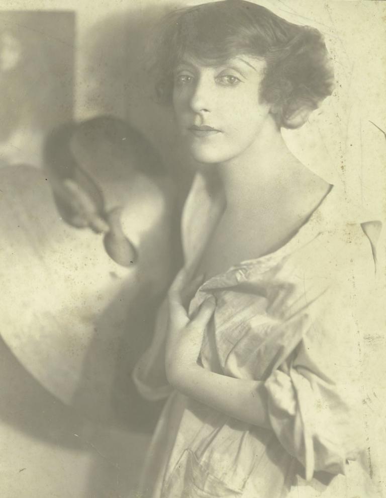 A black and white portrait of Annette Kellerman. She is holding a painting palette with one hand and holding her unbuttoned painter's smock closed with the other.