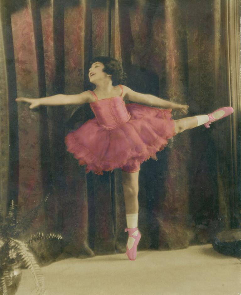 A young girl is wearing ballet tutu and ballet shoes. The photo is a hand-tinted black and white photo.