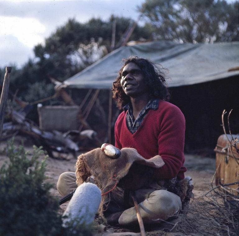 David Gulpilil as Fingerbone Bill sitting down on the outdoor set of Storm Boy holding a fish. There is a boom microphone pointing up at him from the bushes.
