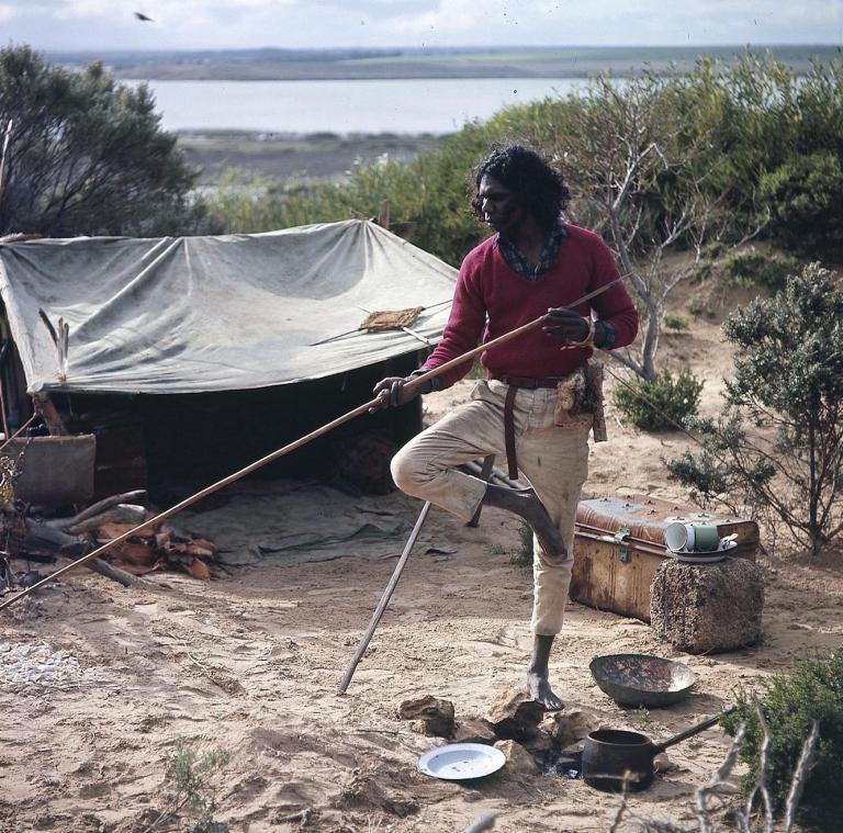 David Gulpilil as Fingerbone Bill standing on one leg holding a spear on the set of his camp near the water from the film Storm Boy