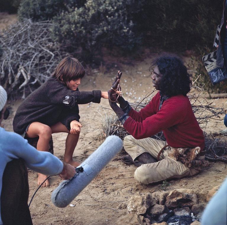 Actors Greg Rowe and David Gulpilil sitting down in the sand, playing with string on the set of Storm Boy. The boom operator with boom and clapper board are visible in the frame