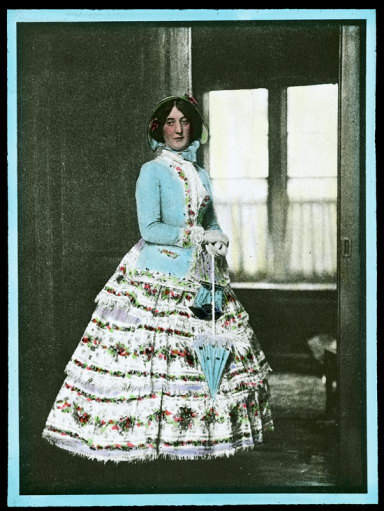 A woman standing near a doorway, wearing a dress from the Victorian era. She has a bonnet, gloves and a parasol. The slide has been hand-coloured.