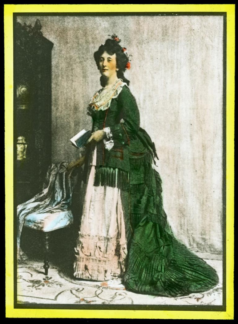 A woman stands in a sitting room holding a book. She is dressed in a late-Victorian era dress with a bustle and has flowers in her hair. The slide is hand-coloured.