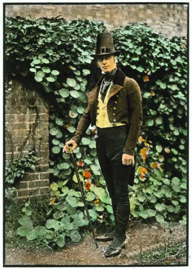 A man stands in front of a garden wall covered in leaves. He's wearing a theatrical costume with hat and waistcoat.