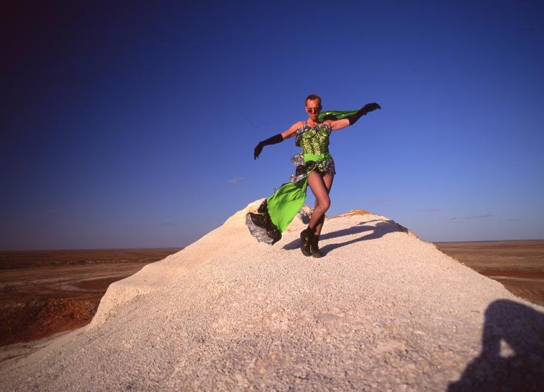 Hugo Weaving in a green dress dancing on a sand dune