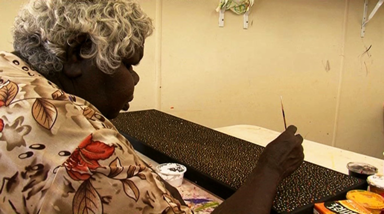 An older Aboriginal woman, pictured from side on, seated at a table painting