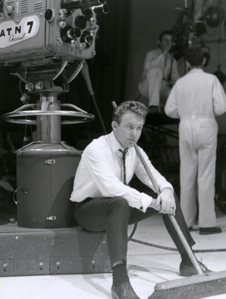 Johnny O'Keefe sitting at the base of a camera holding a broom on the set of 'Sing Sing Sing'