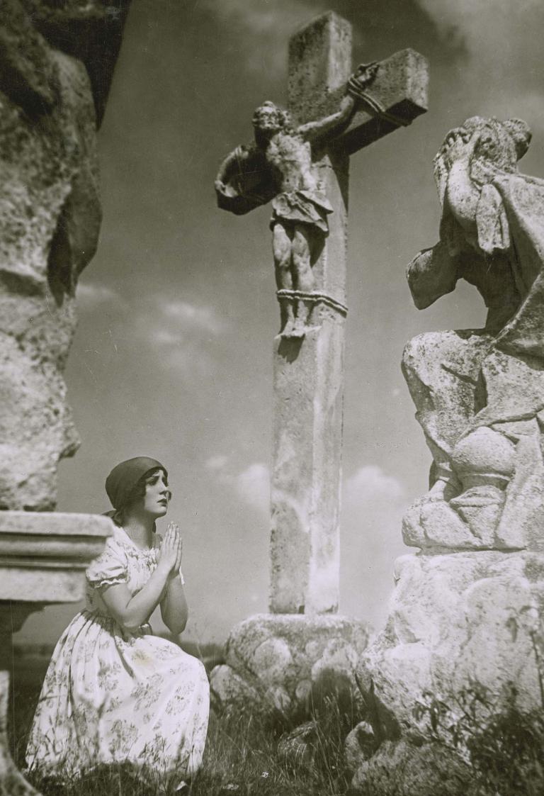 A woman kneels in prayer before a statue of the crucifix in a cemetery. A still from the German film Melody of the Heart, 1929.