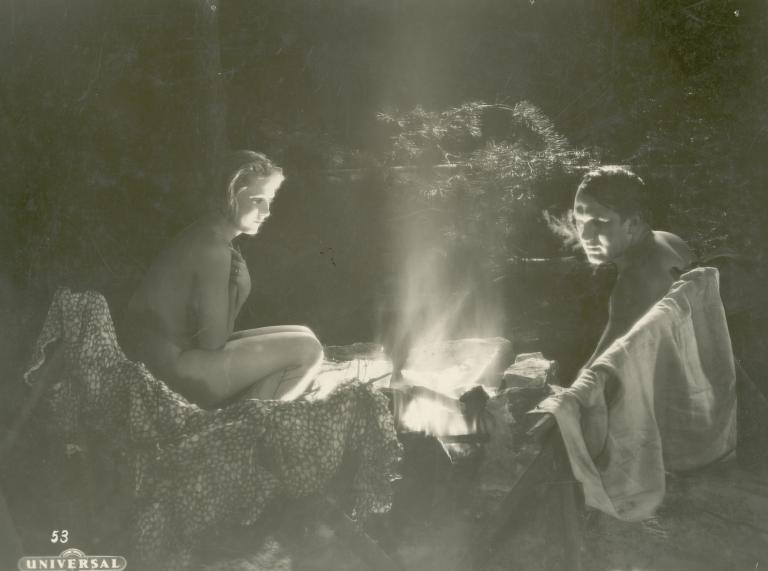 A man and woman sit around an outdoor fire, drying their clothes and keeping warm, in a scene from the German film The Silence in the Forest, 1929.