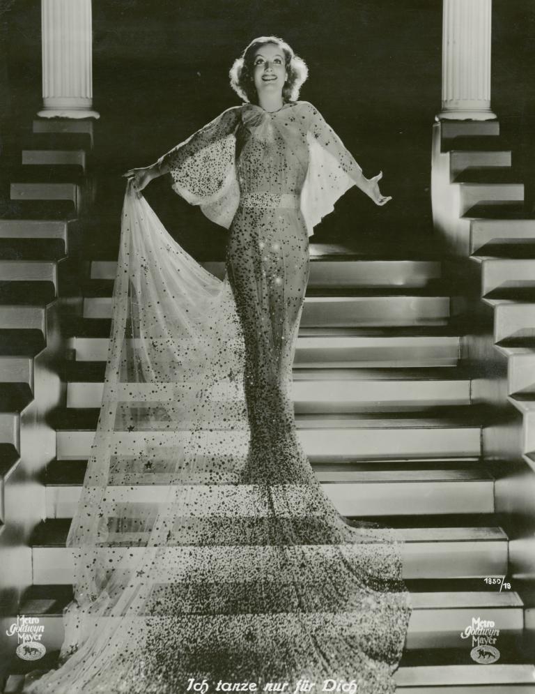 Joan Crawford stands on a staircase, holding the train of her evening dress, in a still from the film Dancing Lady, 1933.