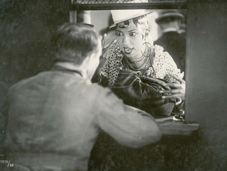 Josephine Baker holds up two fingers to a man at a ticket window in a still from the French film La Sirene des Tropiques, 1927.