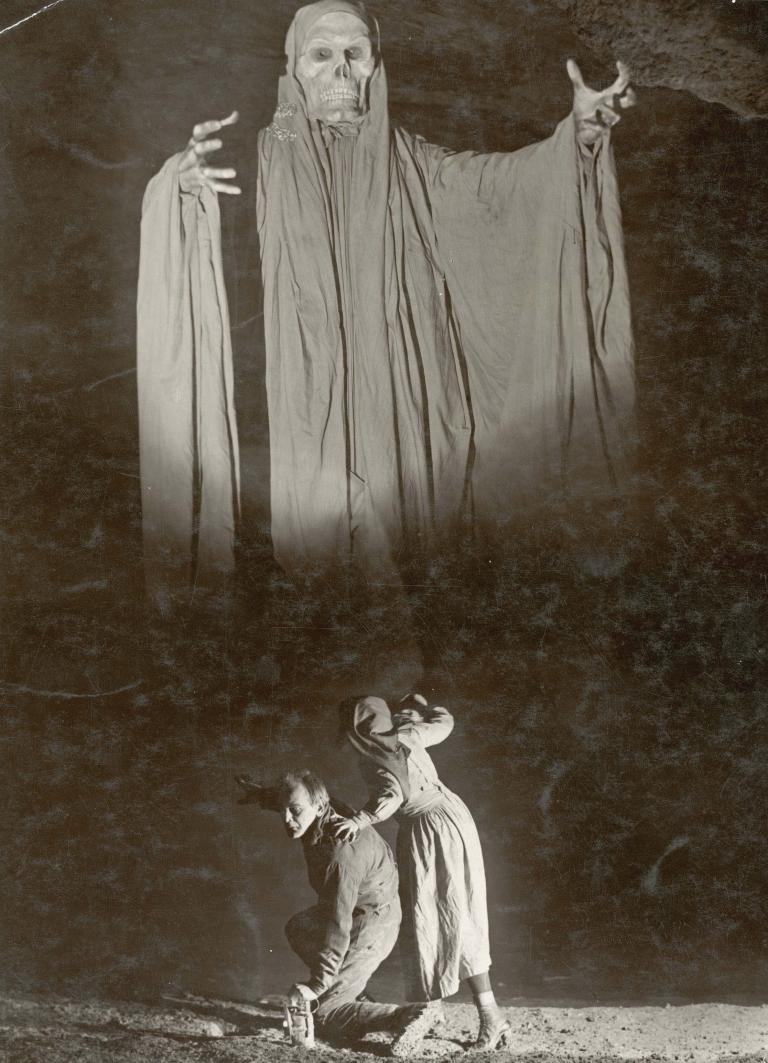 A man and woman cower in the foreground as a menacing skeleton in a cloak looms over them. A still from the Austrian film Irrlichter der Tiefe, 1923.
