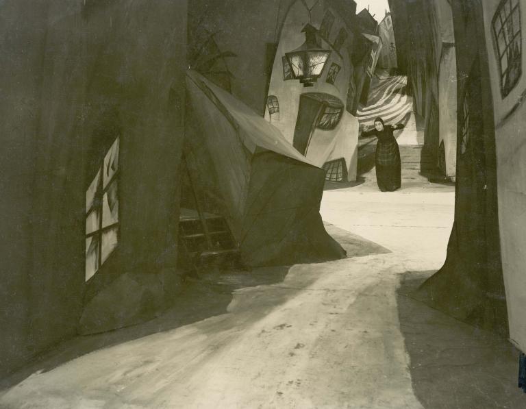 A woman stands in the distance on a surrealistic street with buildings leaning at odd angles. Film still from The Cabinet of Dr Caligari, 1920.
