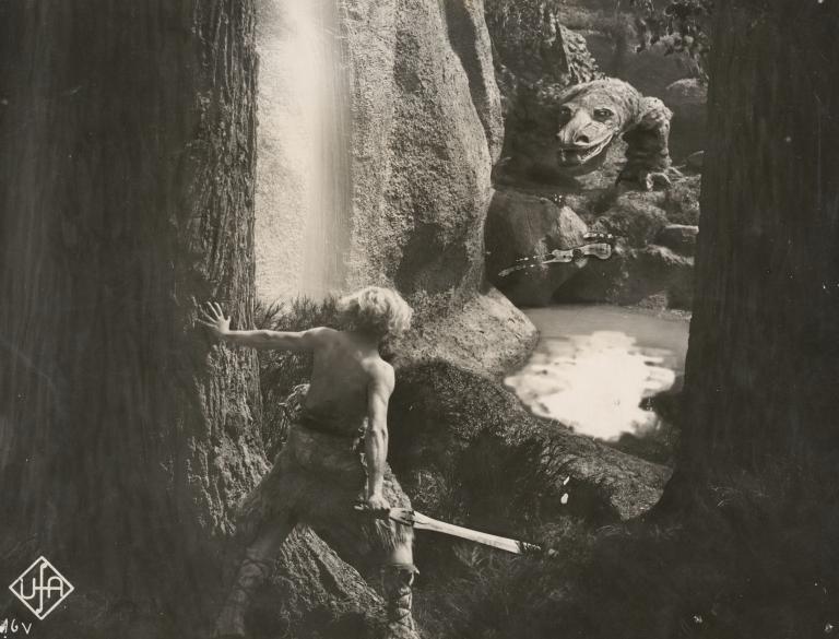 Siegfried is clutching a sword and staring at a crocodile across a pool of water in a still from the film The Nibelungs, 1924.