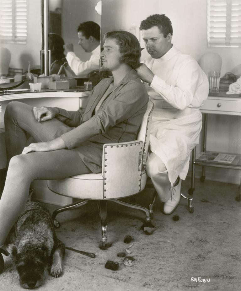 Errol Flynn sits in the make-up chair with a dog at his feet, while a man sits behind him and works on his hair.
