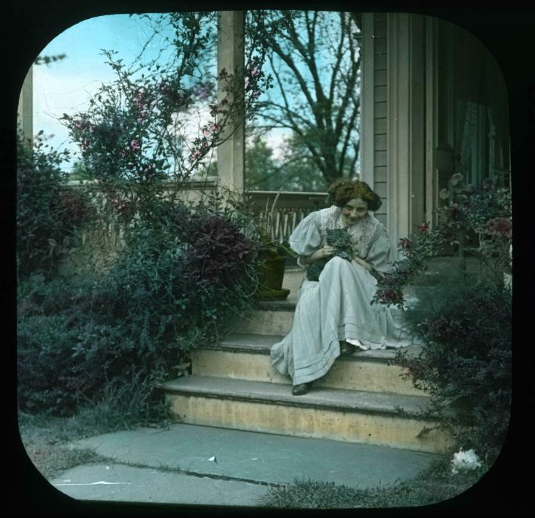 Young woman sitting on some steps in front of a house and holding a grey cat, circa 1906.