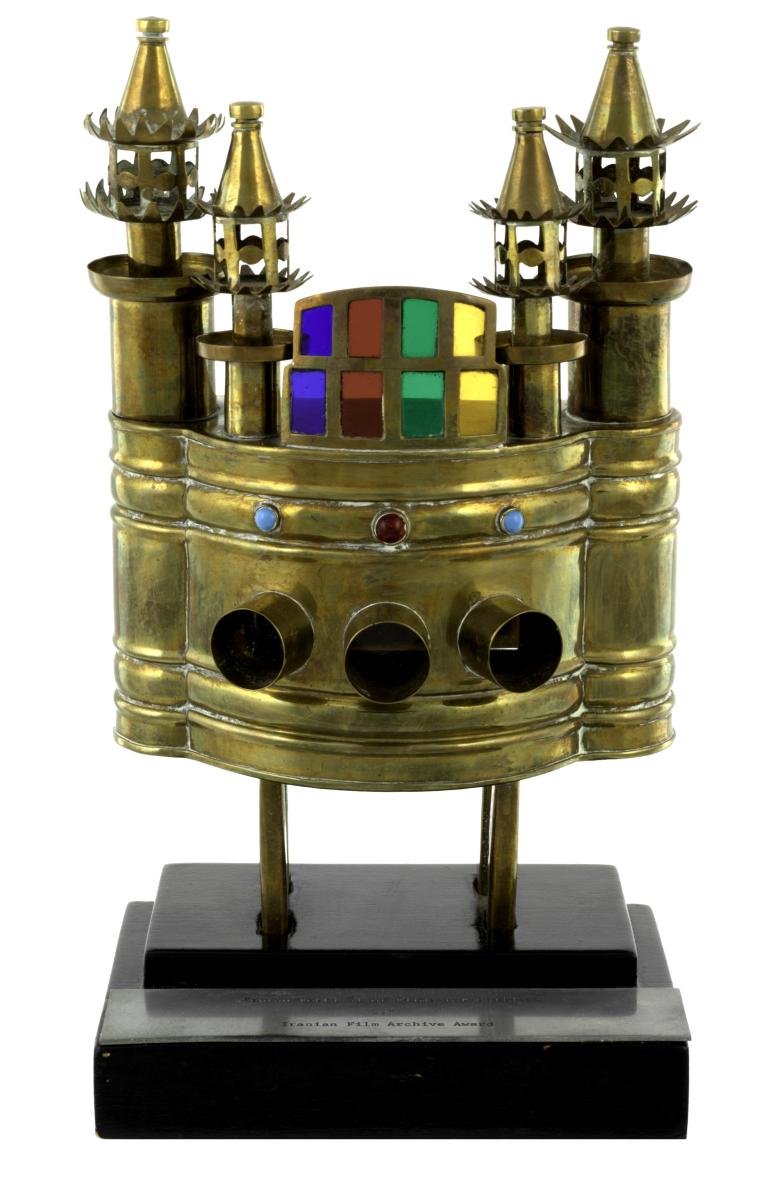 Brass trophy consisting of four legs, a hollow mid section (with door at rear) and three portals at front, four decorative turrets at top and at centre top, a stained glass feature. Also features a painted wooden base with an aluminium plaque