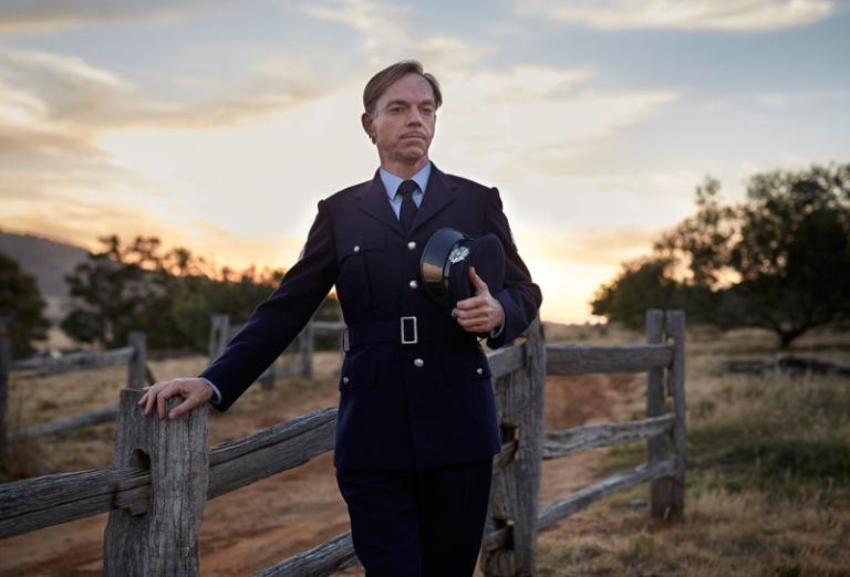 Hugo Weaving as Sergeant Farrat in The Dressmaker. He is standing in uniform alongside a fence.