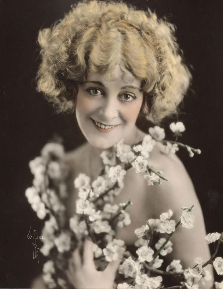 A hand-coloured publicity portrait of actor Vera James. She appears to be naked while holding stems of cherry blossoms in front of her.