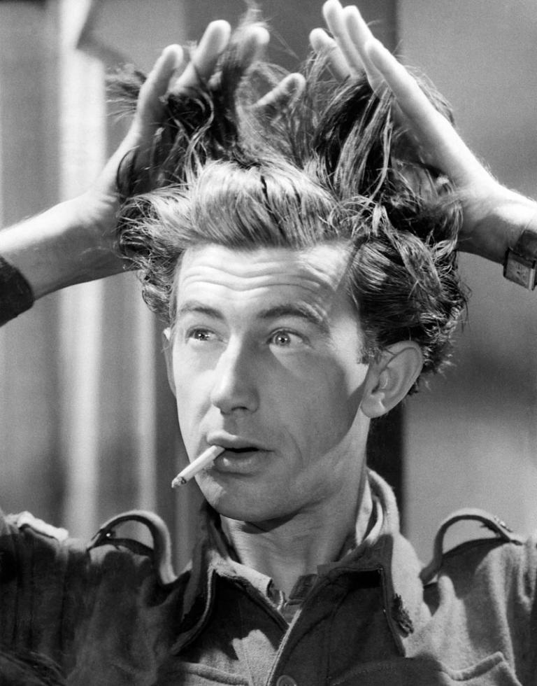 A still of Chips Rafferty holding up his hair with a cigarette hanging out of his mouth.