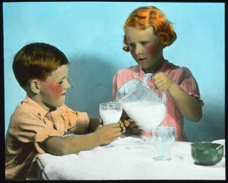 A young girl fills the cup of her smaller companion with milk with a somewhat deadly look in her eye.